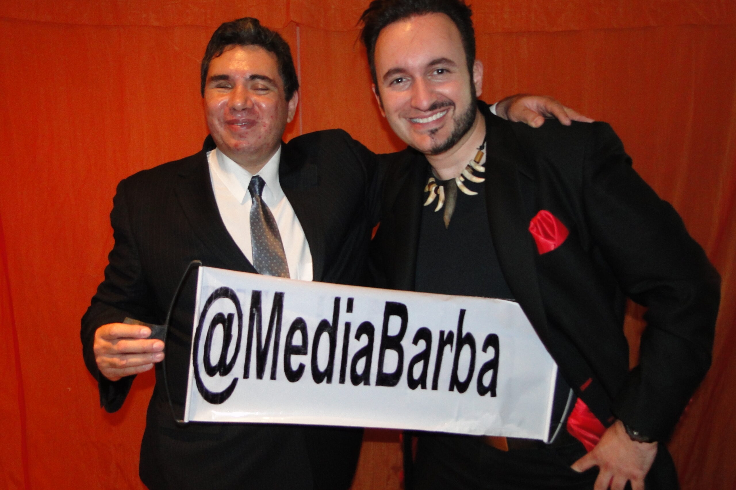 Fotografía de Eagleman junto a MediaBarba en un evento, ambos vestidos de manera formal, sosteniendo un cartel con el nombre “@MediaBarba”.